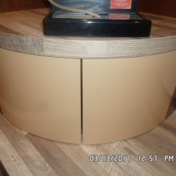 F.H.P.U.DREWMAK - Zaokrąglona szafka z beżowymi, lakierowanymi frontami i blatem imitującym drewno. Na blacie stoi czarny ekspres do kawy. Widoczna podłoga z paneli.