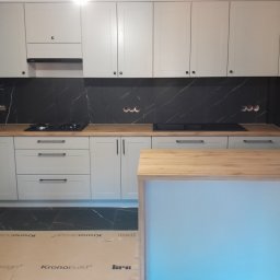F.H.P.U.DREWMAK - Jasnoszara kuchnia na wymiar z drewnianym blatem i czarnym marmurowym backsplashiem. Widoczna wyspa kuchenna z drewnianym blatem i niebieskim podświetleniem.