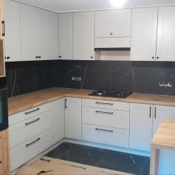 F.H.P.U.DREWMAK - Jasna kuchnia z drewnianym blatem i czarnymi płytkami imitującymi marmur. Widoczna zabudowa piekarnika i płyta gazowa. Minimalistyczne uchwyty.