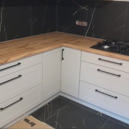 F.H.P.U.DREWMAK - Jasnoszara zabudowa kuchenna z drewnianym blatem i czarnymi uchwytami na tle czarnych płytek imitujących marmur. Widoczna płyta gazowa.