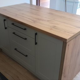 F.H.P.U.DREWMAK - Szafka z blatem drewnianym, szare fronty, czarne uchwyty, widok z boku, na podłodze panele, w tle fragment podłogi wyłożonej czarnym gresem.