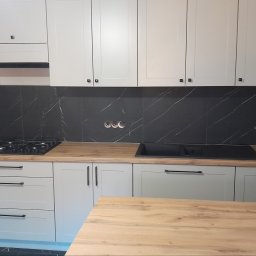 F.H.P.U.DREWMAK - Jasnoszara zabudowa kuchenna z drewnianym blatem i czarnym panelem ściennym imitującym marmur. Widoczne gniazdka elektryczne i płyta grzewcza.