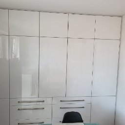 F.H.P.U.DREWMAK - Biała, lakierowana szafa wnękowa z metalowymi uchwytami, podzielona na segmenty, sięgająca sufitu. Widoczny fragment biurka i krzesła.