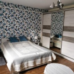F.H.P.U.DREWMAK - Sypialnia z łóżkiem, szafą przesuwną z lustrem i wzorzystą tapetą w odcieniach błękitu i szarości. Widoczny fragment podłogi z paneli.