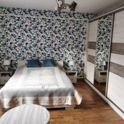 F.H.P.U.DREWMAK - Sypialnia z dużą szafą przesuwną z lustrem i białymi frontami z szarymi wstawkami. Na łóżku dwie niebieskie poduszki. Na ścianie wzorzysta tapeta.