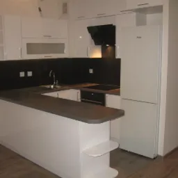 Nowoczesna kuchnia z białymi, lakierowanymi frontami, czarnym panelem nad blatem i szarym blatem, widok na zabudowę z wyspą i lodówką Amica.