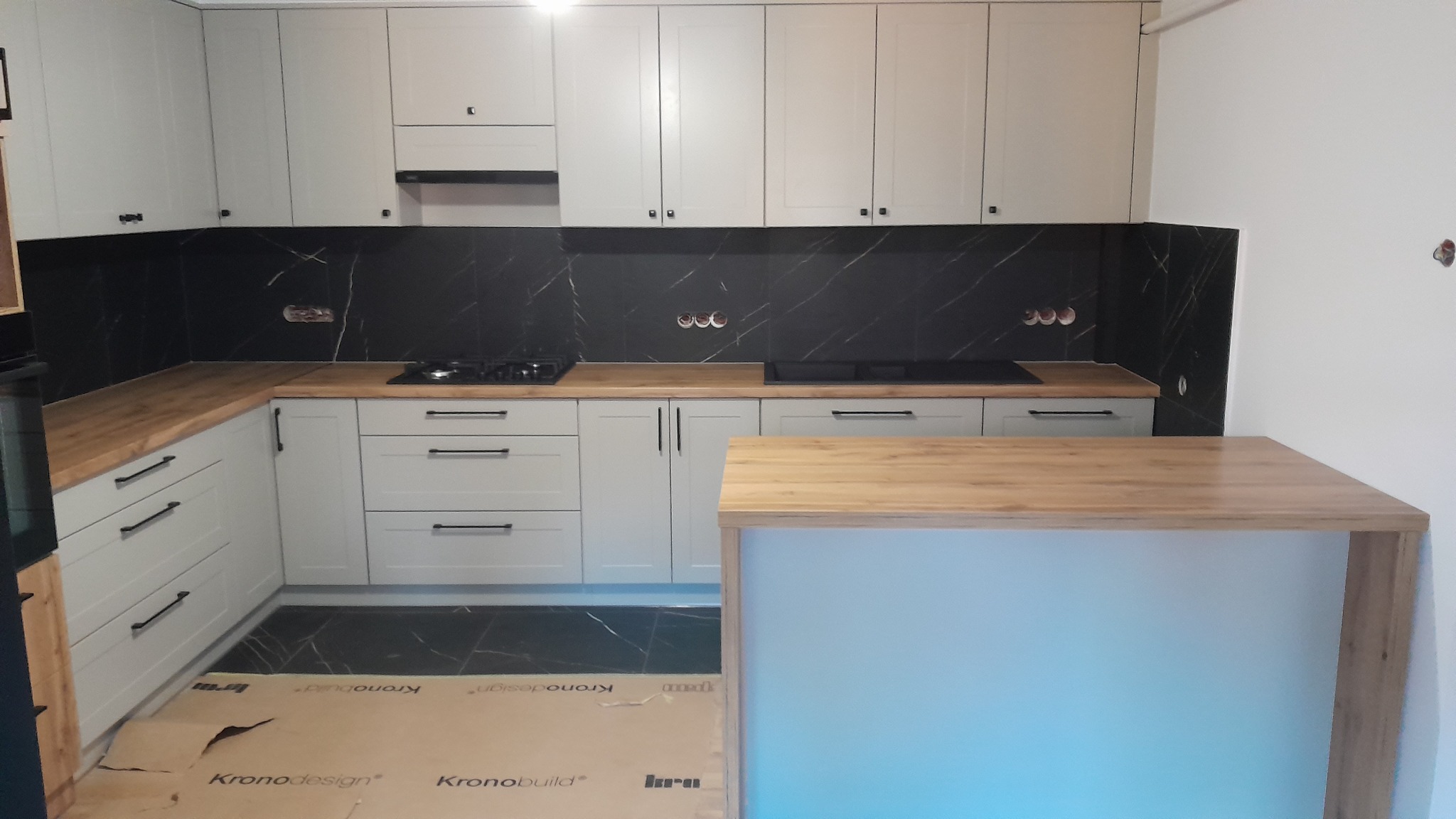 Jasnoszara kuchnia na wymiar z drewnianym blatem i czarnym marmurowym backsplashiem. Widoczna wyspa kuchenna z drewnianym blatem i niebieskim podświetleniem.