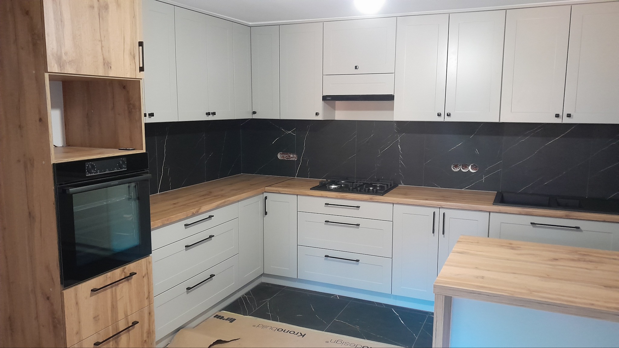 Jasna kuchnia z drewnianym blatem i czarnymi płytkami imitującymi marmur. Widoczna zabudowa piekarnika i płyta gazowa. Minimalistyczne uchwyty.