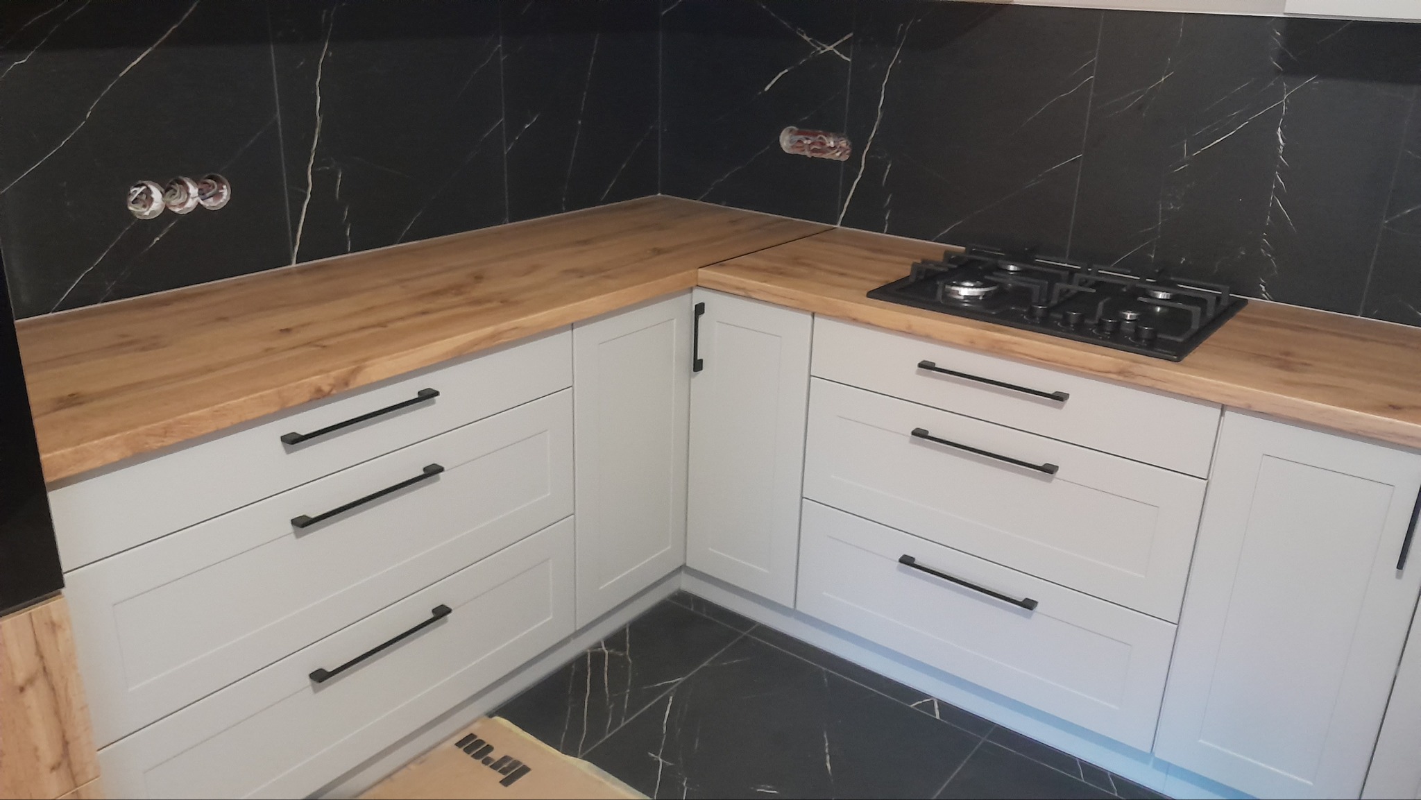 Jasnoszara zabudowa kuchenna z drewnianym blatem i czarnymi uchwytami na tle czarnych płytek imitujących marmur. Widoczna płyta gazowa.