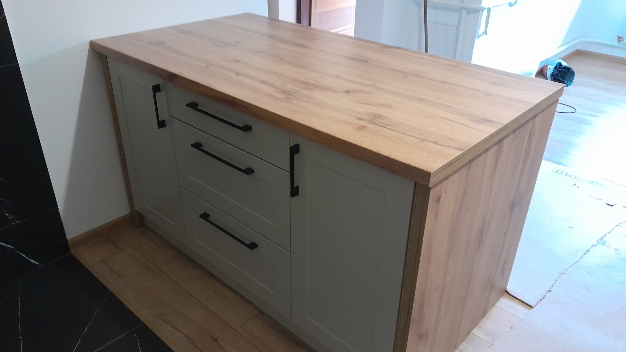 Szafka z blatem drewnianym, szare fronty, czarne uchwyty, widok z boku, na podłodze panele, w tle fragment podłogi wyłożonej czarnym gresem.