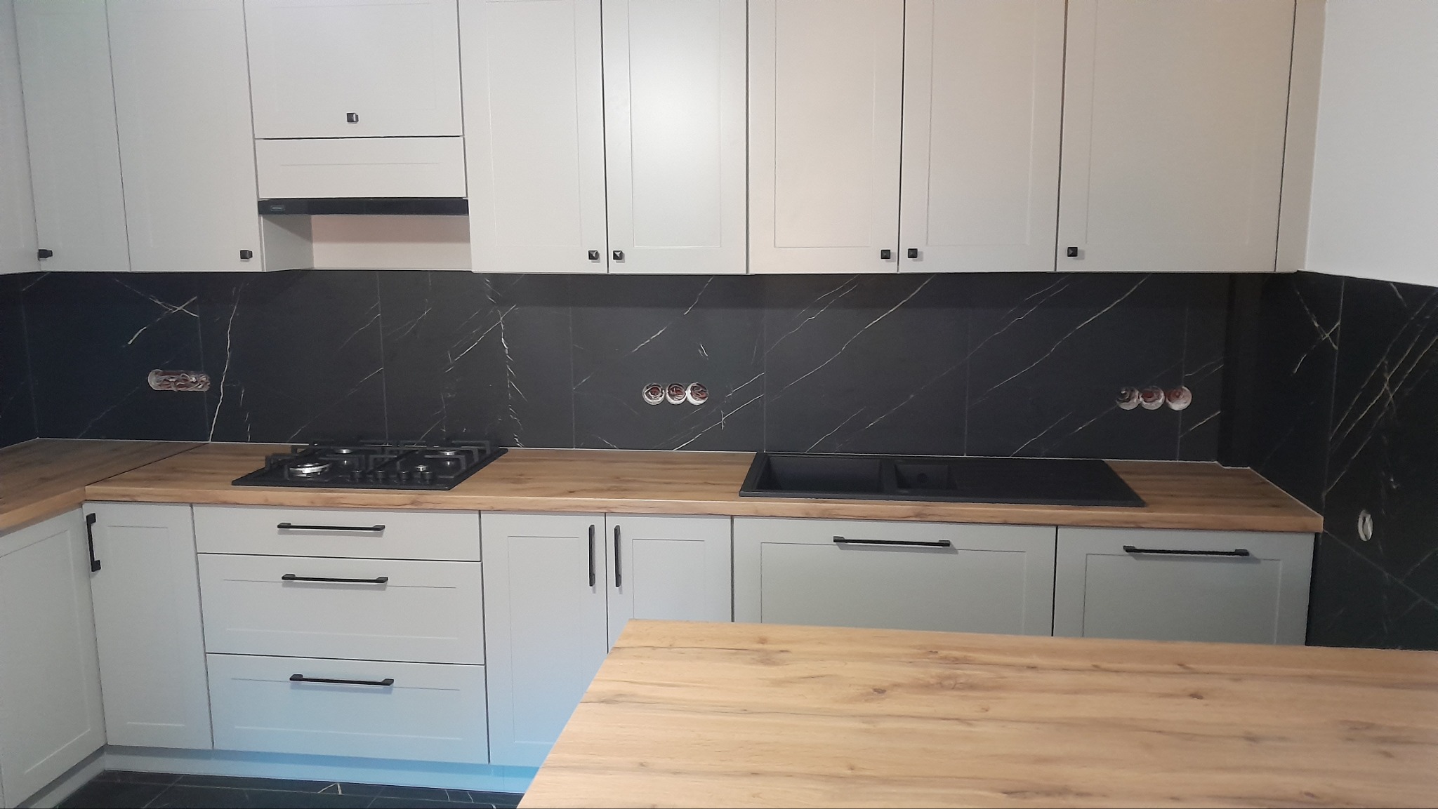 Jasnoszara zabudowa kuchenna z drewnianym blatem i czarnym panelem ściennym imitującym marmur. Widoczne gniazdka elektryczne i płyta grzewcza.