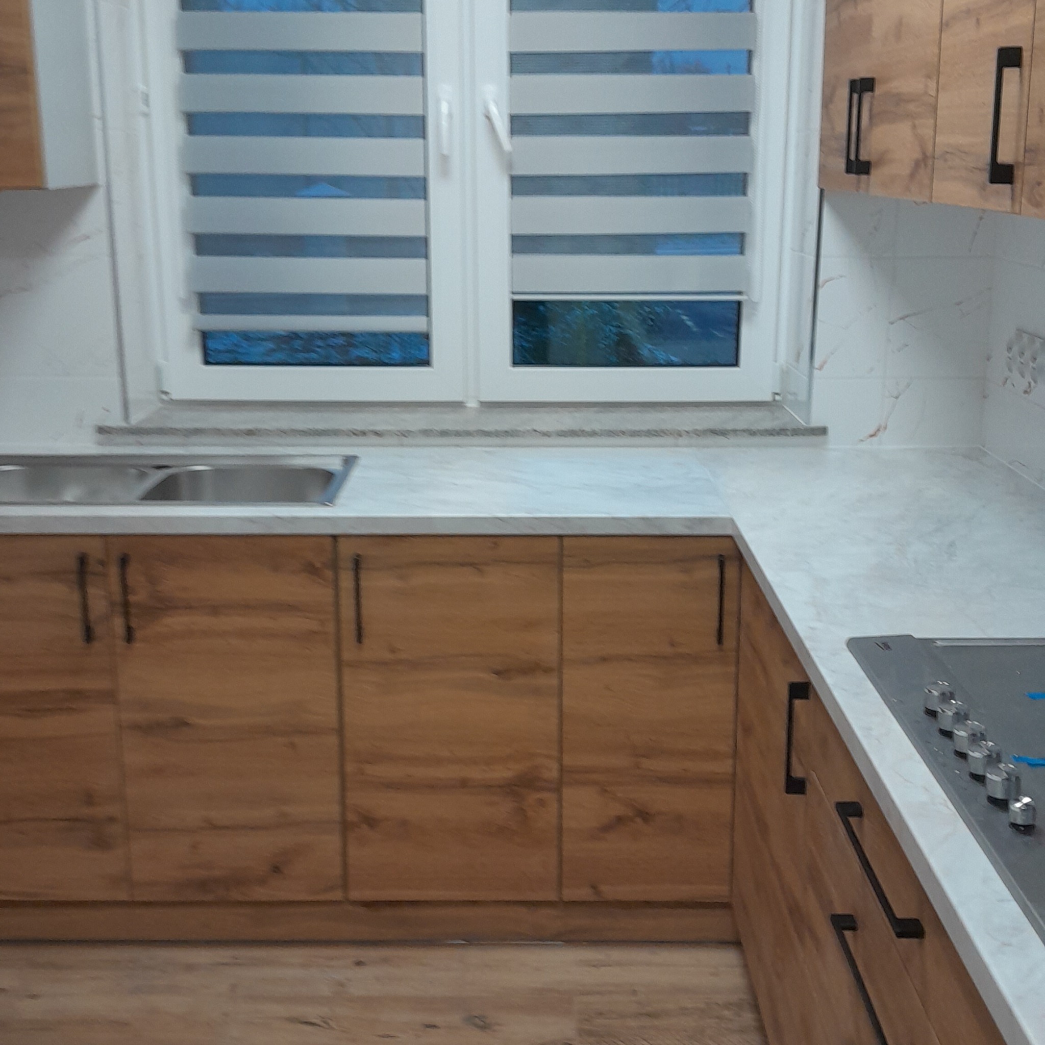 Kuchenna zabudowa z drewnianymi frontami i czarnymi uchwytami, jasny blat, okno z żaluzjami w tle. Ujęcie z niskiego kąta, widoczna podłoga.