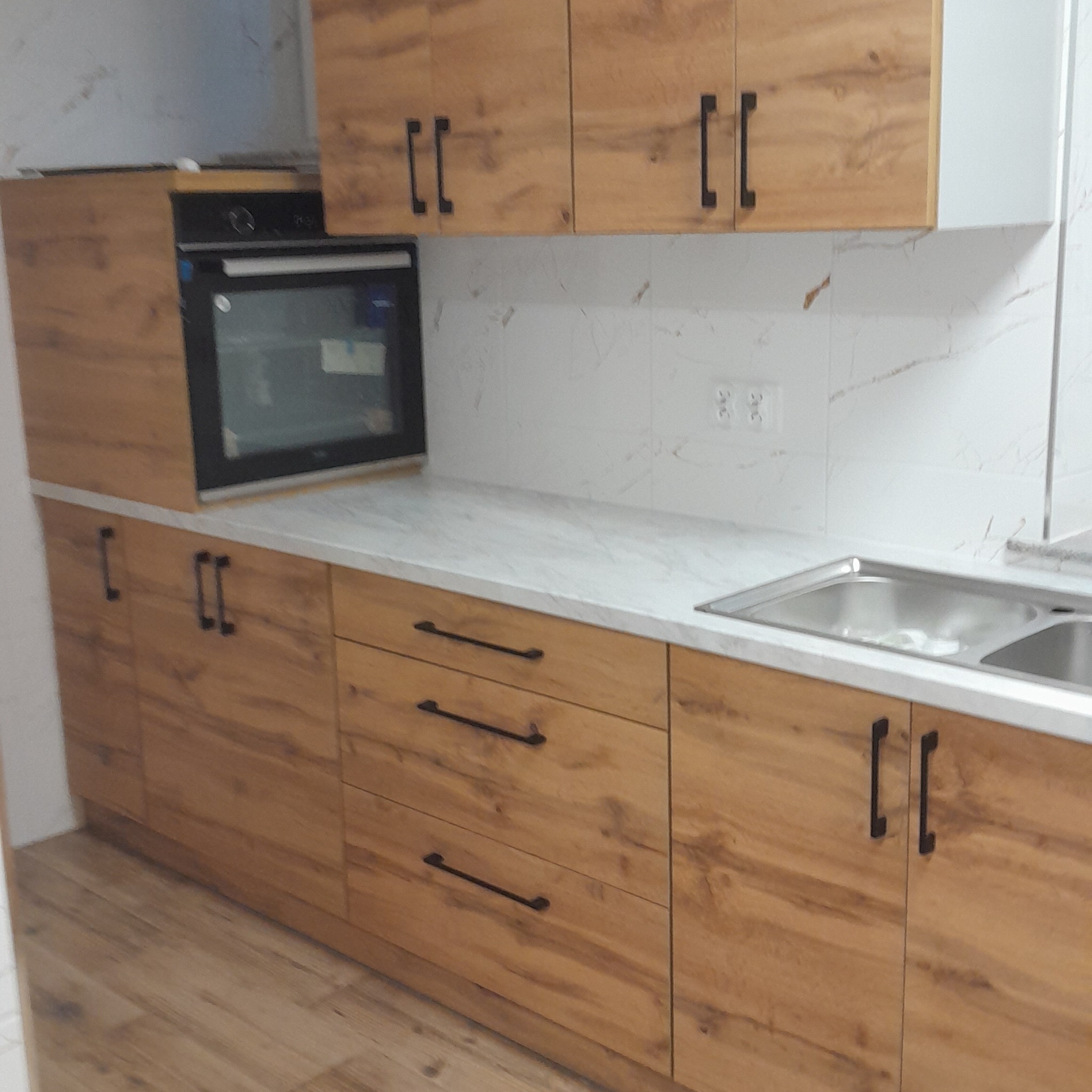 Nowoczesna kuchnia z drewnianymi frontami i czarnymi uchwytami. Blat imitujący marmur, zlew dwukomorowy. Widoczny piekarnik w zabudowie.