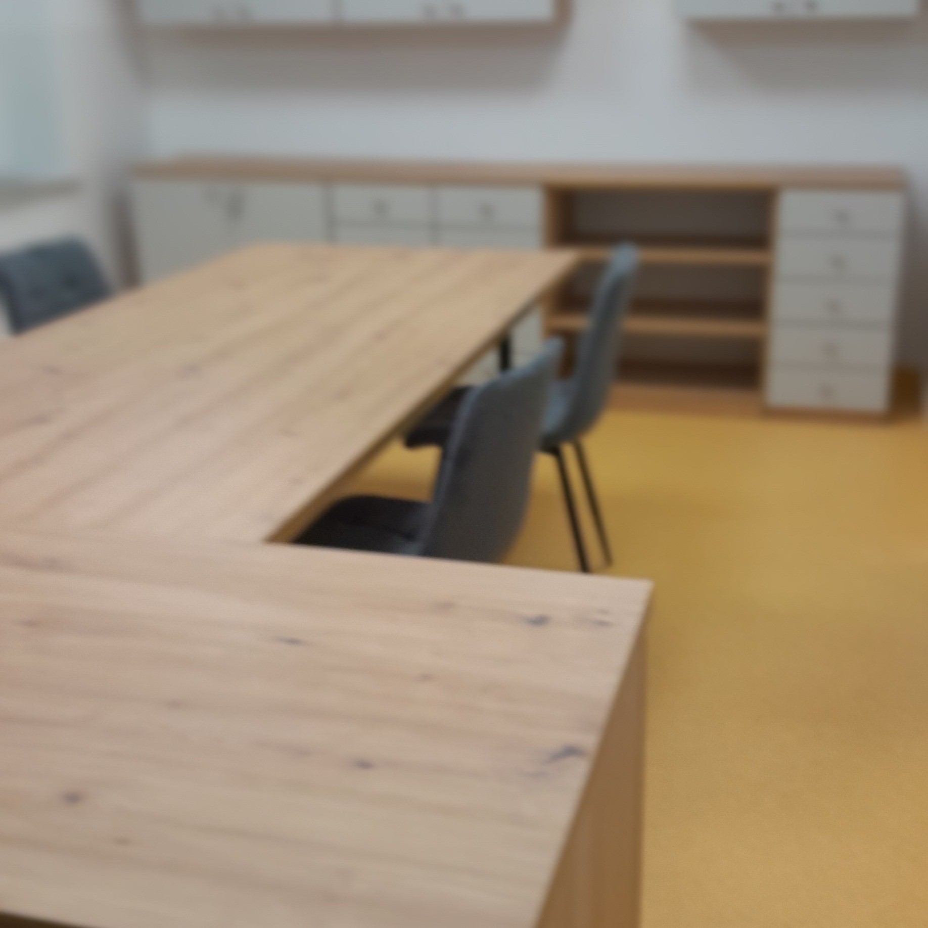 Jasny, drewniany blat biurka w kształcie litery U z szarymi krzesłami, na tle białych szafek z drewnianymi akcentami i żółtej podłogi. Ujęcie z bliska, lekko rozmyte.