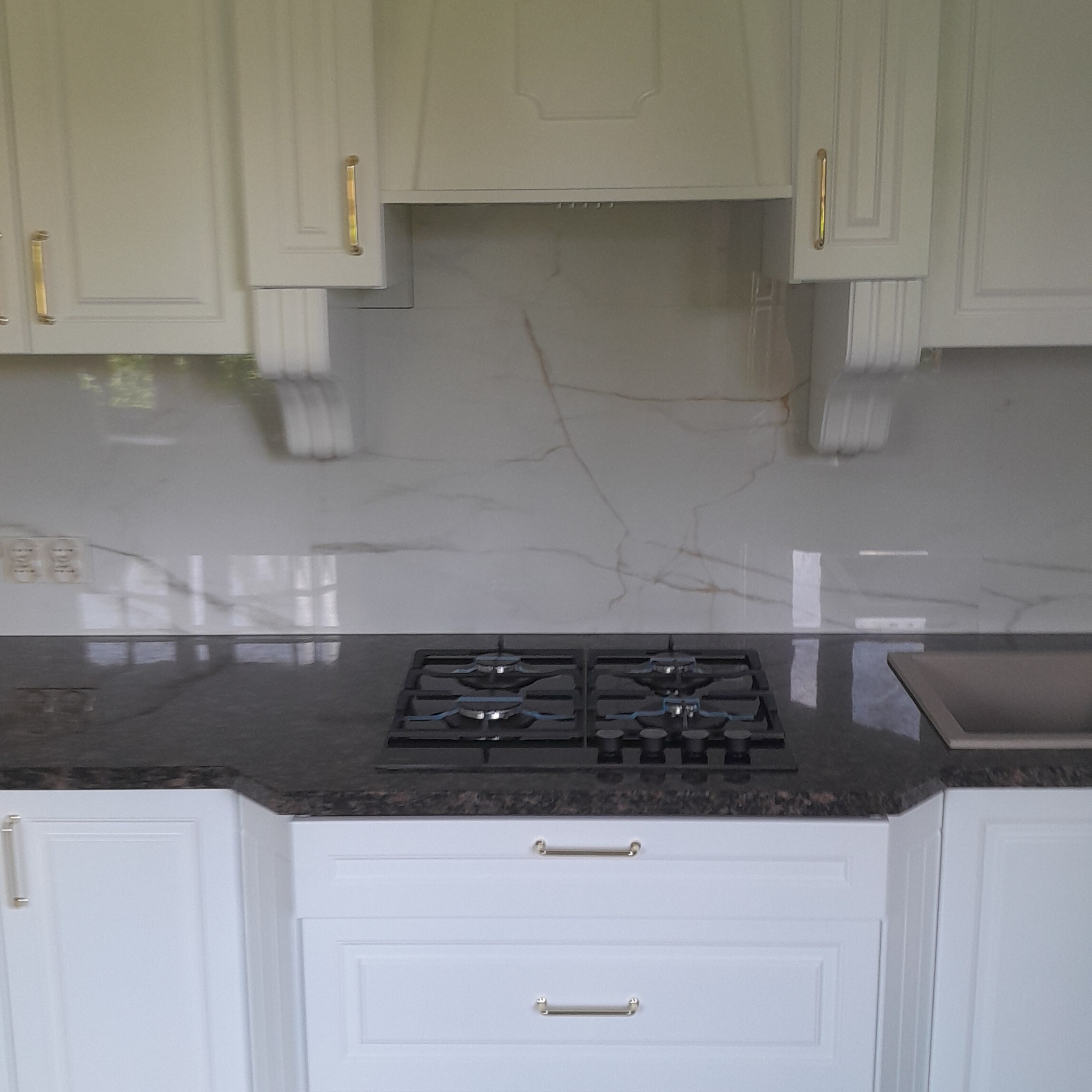 Biała kuchnia z frezowanymi frontami, czarnym blatem i płytą gazową. Złote uchwyty dodają elegancji. Marmurowy wzór na ścianie.