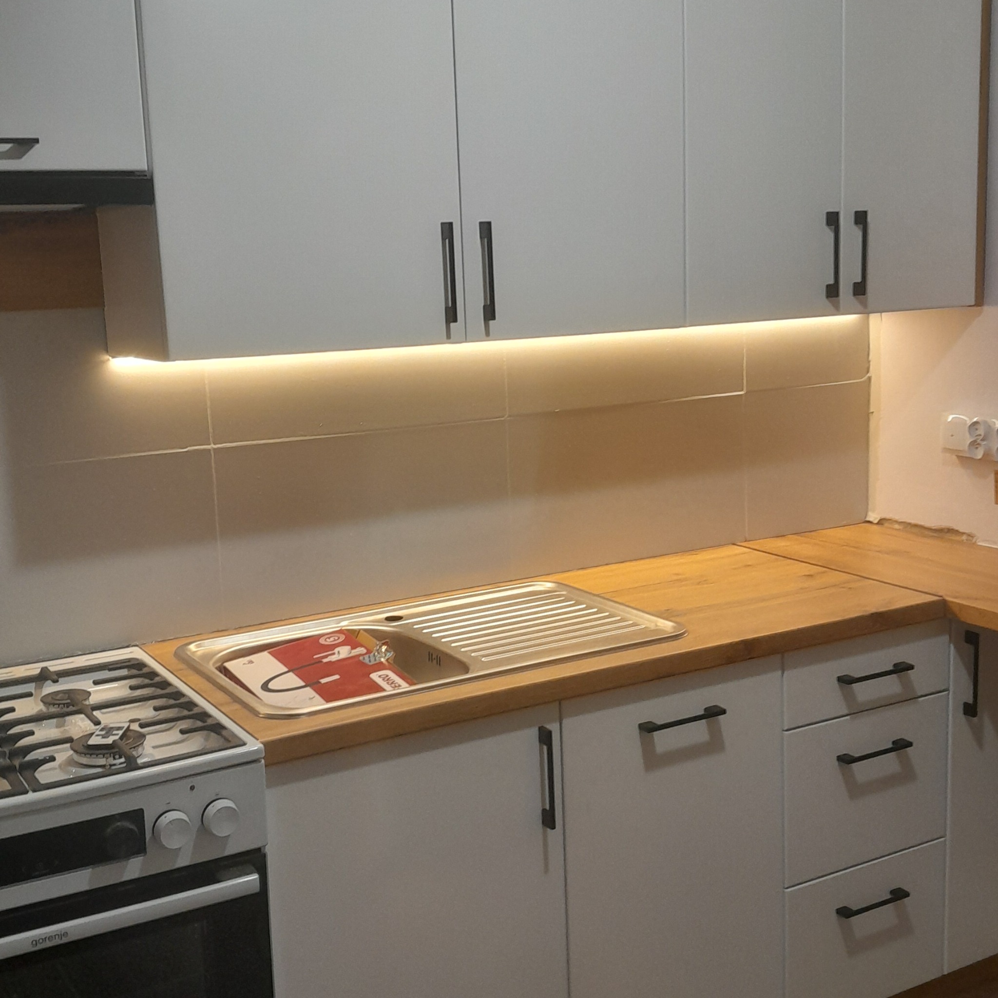 Jasna kuchnia z drewnianym blatem i oświetleniem LED pod szafkami. Widoczny zlew z ociekaczem, płyta gazowa i minimalistyczne, czarne uchwyty.