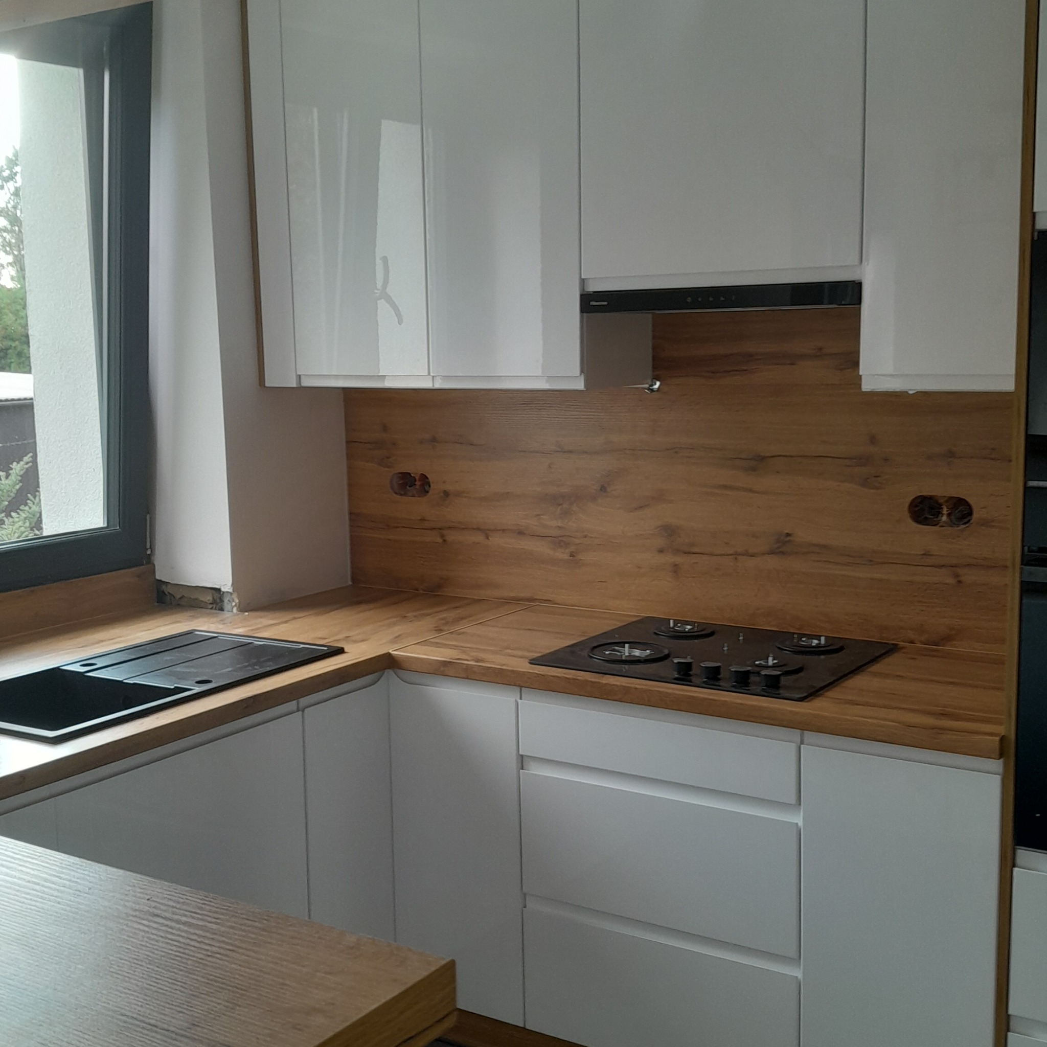 Białe szafki kuchenne z drewnianym blatem i okładziną, czarny zlew i płyta grzewcza. Widoczne gniazdka elektryczne w okładzinie.