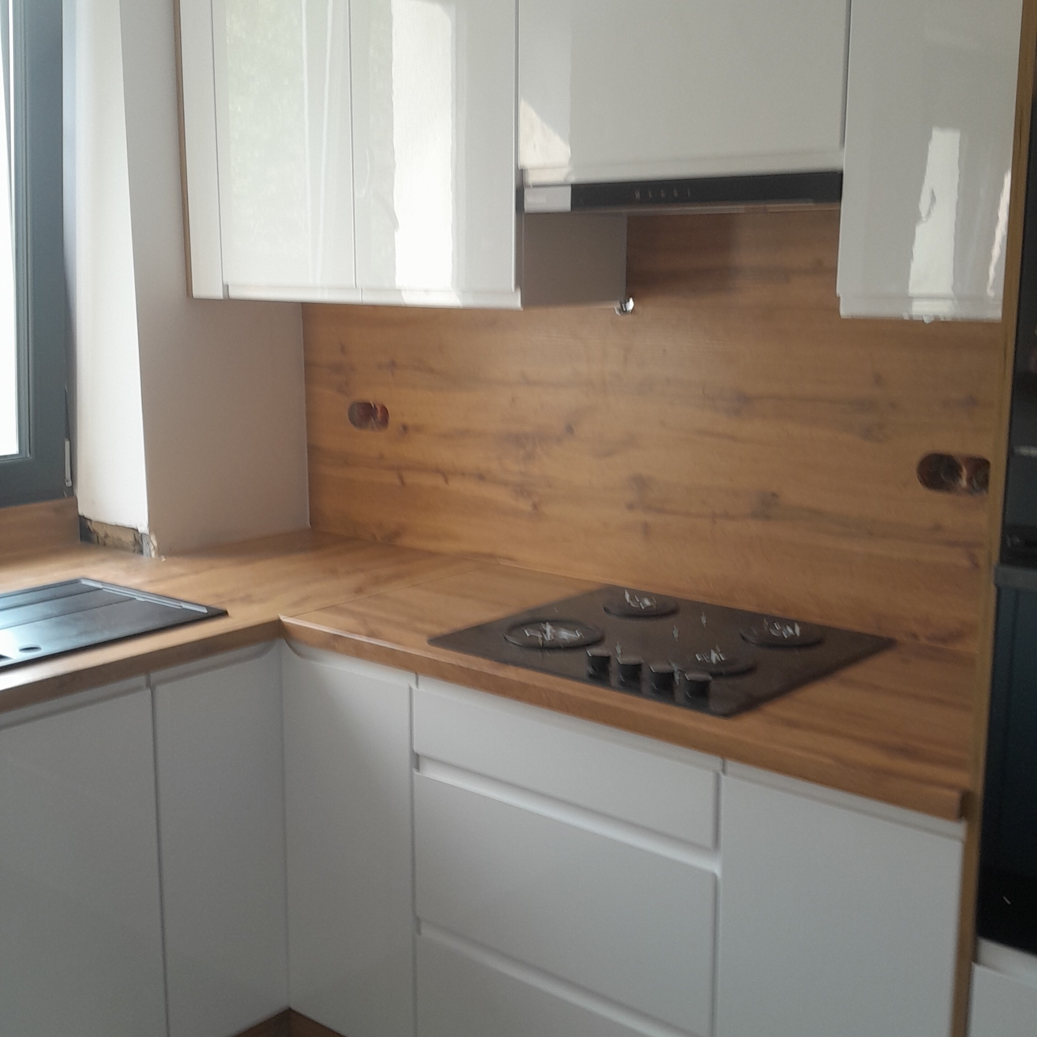 Białe szafki kuchenne z drewnianym blatem i panelem ściennym. Na blacie płyta grzewcza. Widoczne gniazdka elektryczne w panelu. Minimalistyczny design.