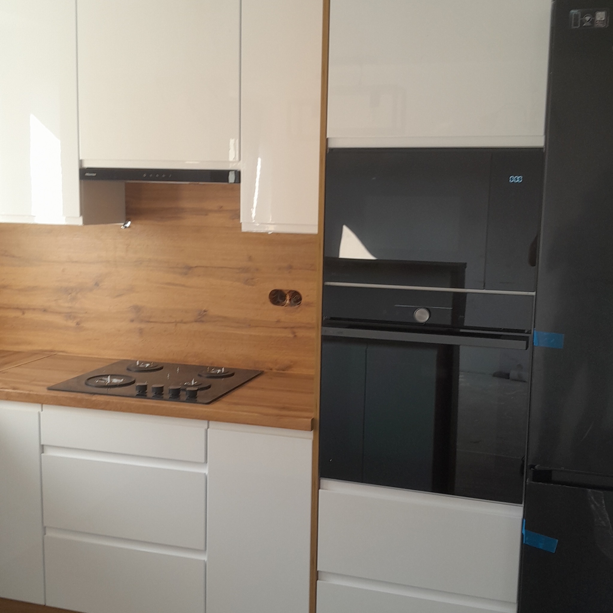 Biała zabudowa kuchenna z drewnianym blatem i płytą grzewczą. W tle piekarnik i lodówka w czarnym kolorze. Minimalistyczny design.