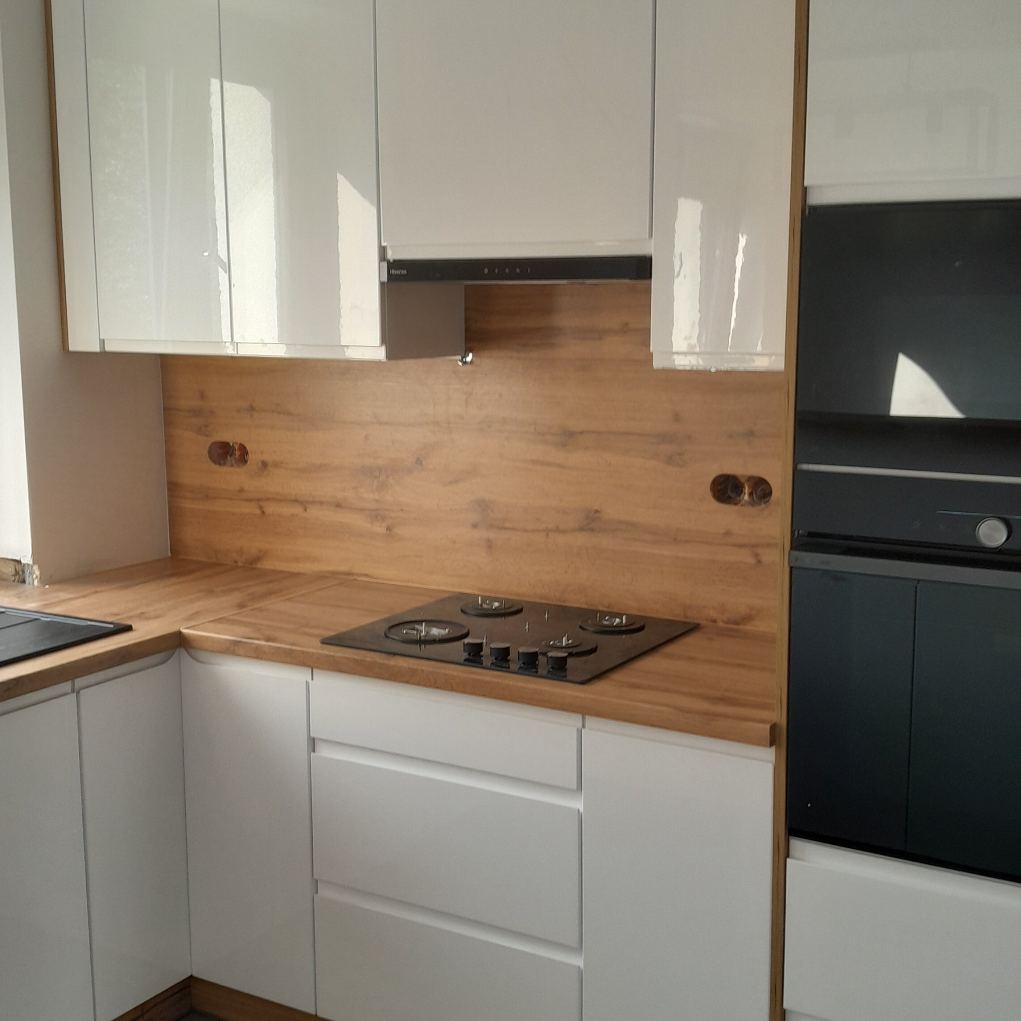 Biała kuchnia na wymiar z drewnianym blatem i płytą grzewczą. Widoczne gniazdka elektryczne w drewnianej ściance nad blatem. Nowoczesny design.