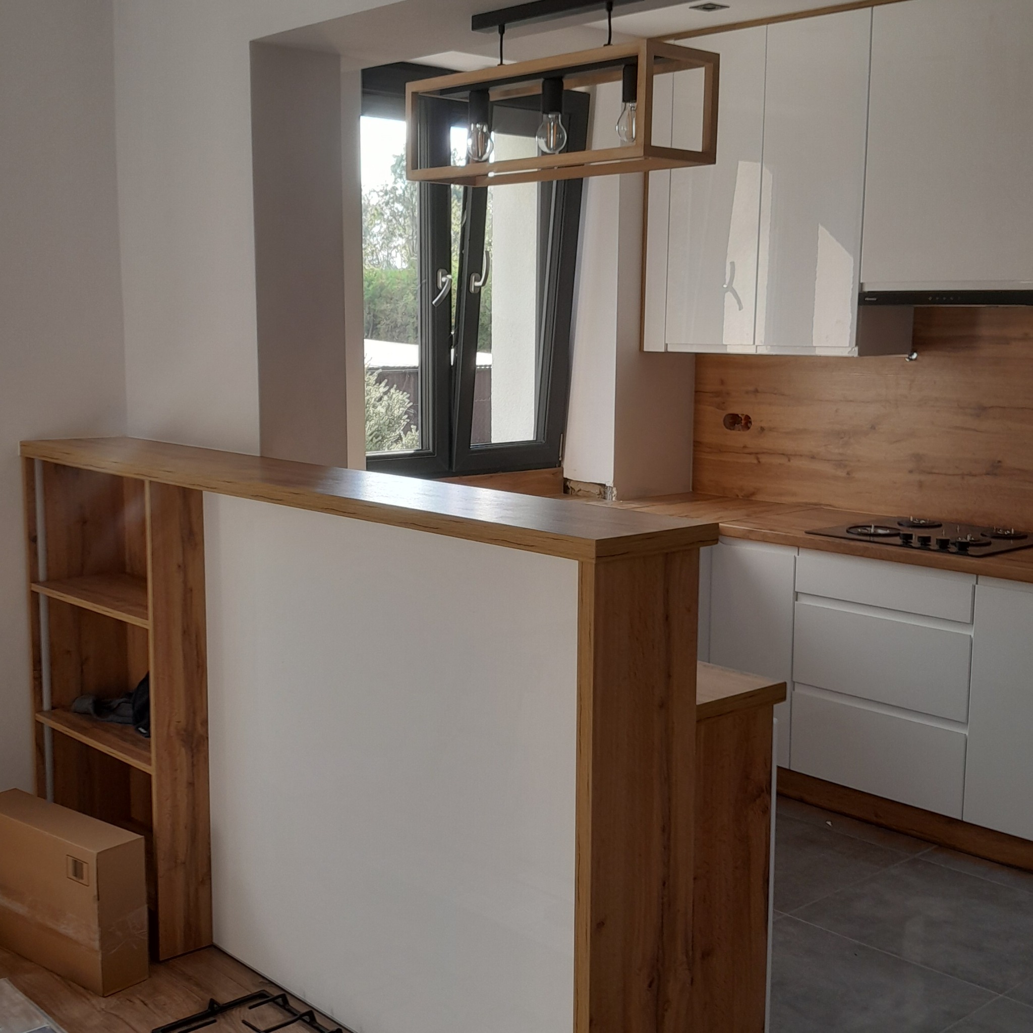 Nowoczesna kuchnia z białymi frontami i drewnianym blatem, oświetlona lampą w stylu industrialnym. Widoczny barek z półkami i okno z widokiem na zieleń.