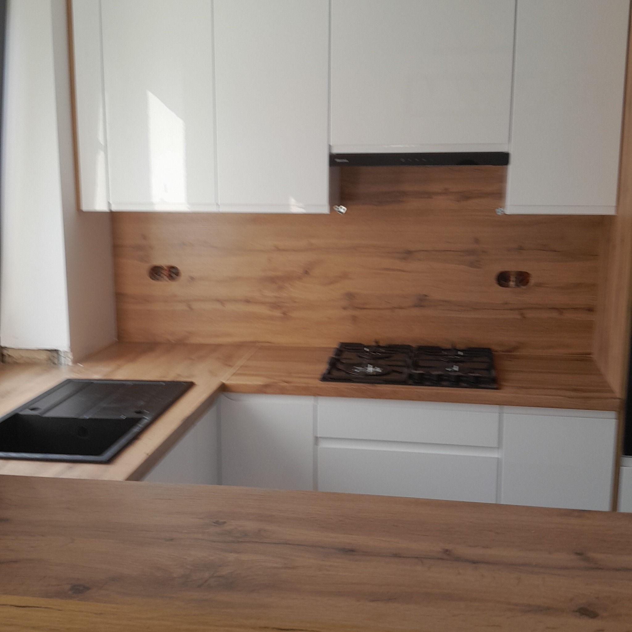 Nowoczesna kuchnia z białymi szafkami i drewnianym blatem. Widoczny zlew, płyta gazowa i fragment blatu wyspowego. Ciepłe oświetlenie podkreśla naturalny wygląd drewna.