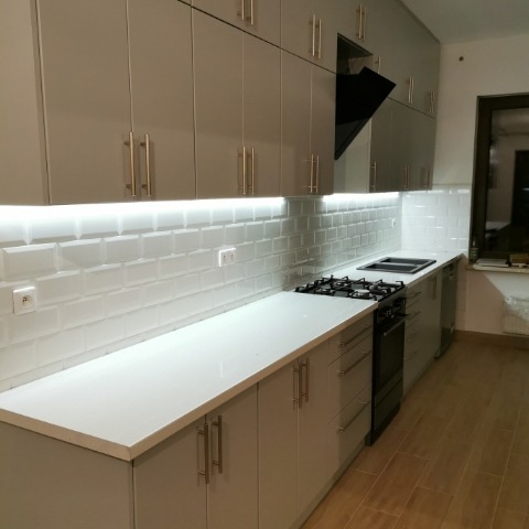 Jasnoszara zabudowa kuchenna z białym blatem i podświetleniem LED w Kolbuszowej. Płytki ścienne typu metro i złote uchwyty dodają elegancji.