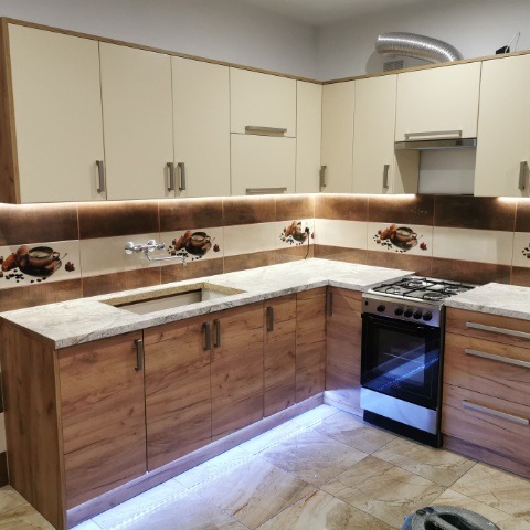 Kremowo-brązowa kuchnia narożna z oświetleniem LED podszafkowym i płytkami z motywem kawy. Jasny blat i fronty szafek dolnych z wzorem drewna.