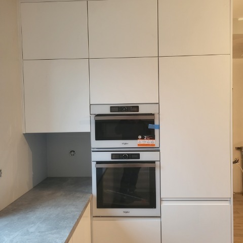 Biała zabudowa kuchenna z piekarnikiem i mikrofalówką Whirlpool. Minimalistyczny design, gładkie fronty szafek, szary blat. Widoczne gniazdko elektryczne.
