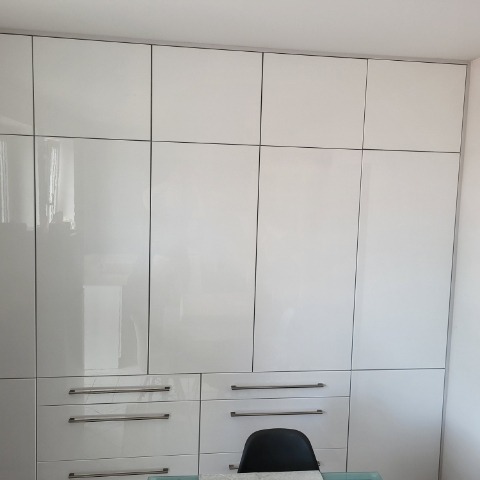 Biała, lakierowana szafa wnękowa z metalowymi uchwytami, podzielona na segmenty, sięgająca sufitu. Widoczny fragment biurka i krzesła.