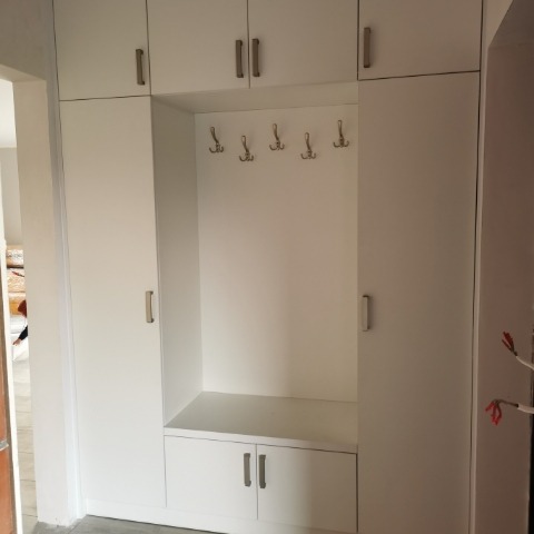 Biała szafa wnękowa z siedziskiem i wieszakami, minimalistyczny design, jasne wnętrze, widok z przodu, proste uchwyty, funkcjonalne rozwiązanie do przedpokoju.