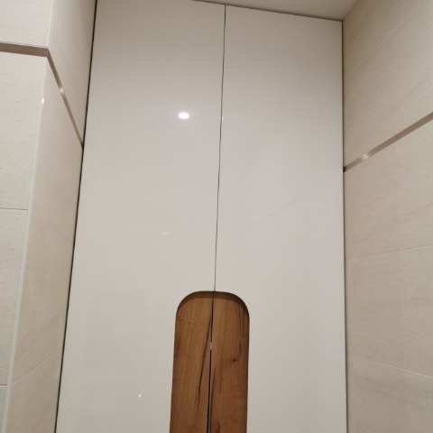 Biała szafa wnękowa z drewnianym akcentem, wkomponowana w łazienkę z beżowymi płytkami. Minimalistyczny design, gładkie fronty, subtelne oświetlenie.