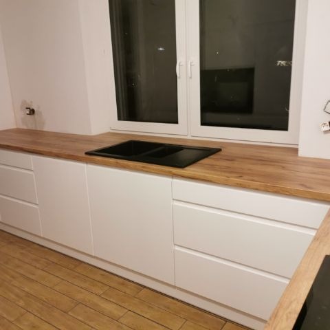 Białe szafki kuchenne z drewnianym blatem i czarnym zlewem pod oknem. Widoczna drewniana podłoga. Minimalistyczny design.