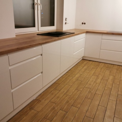 Biała, narożna kuchnia z drewnianym blatem i płytą grzewczą, widok na podłogę z drewnopodobnych płytek. Minimalistyczny design, jasne wnętrze.