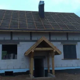 Surowy stan domu z białych bloczków, widoczne przygotowania pod dach, drewniana konstrukcja nad wejściem, okna zabezpieczone folią, komin owinięty folią, teren wokół w trakcie przygotowania.