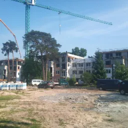 Wczesny etap budowy kilku budynków wielorodzinnych, widoczne konstrukcje w stanie surowym, dźwigi budowlane, mieszalnik betonu, materiały budowlane na paletach i zaparkowane samochody.