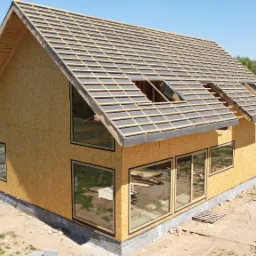 Surowy stan domu z drewnianą konstrukcją szkieletową i oknami, widoczne ułożone elementy dachu i przygotowane fundamenty, w tle drugi dom z panelami solarnymi.