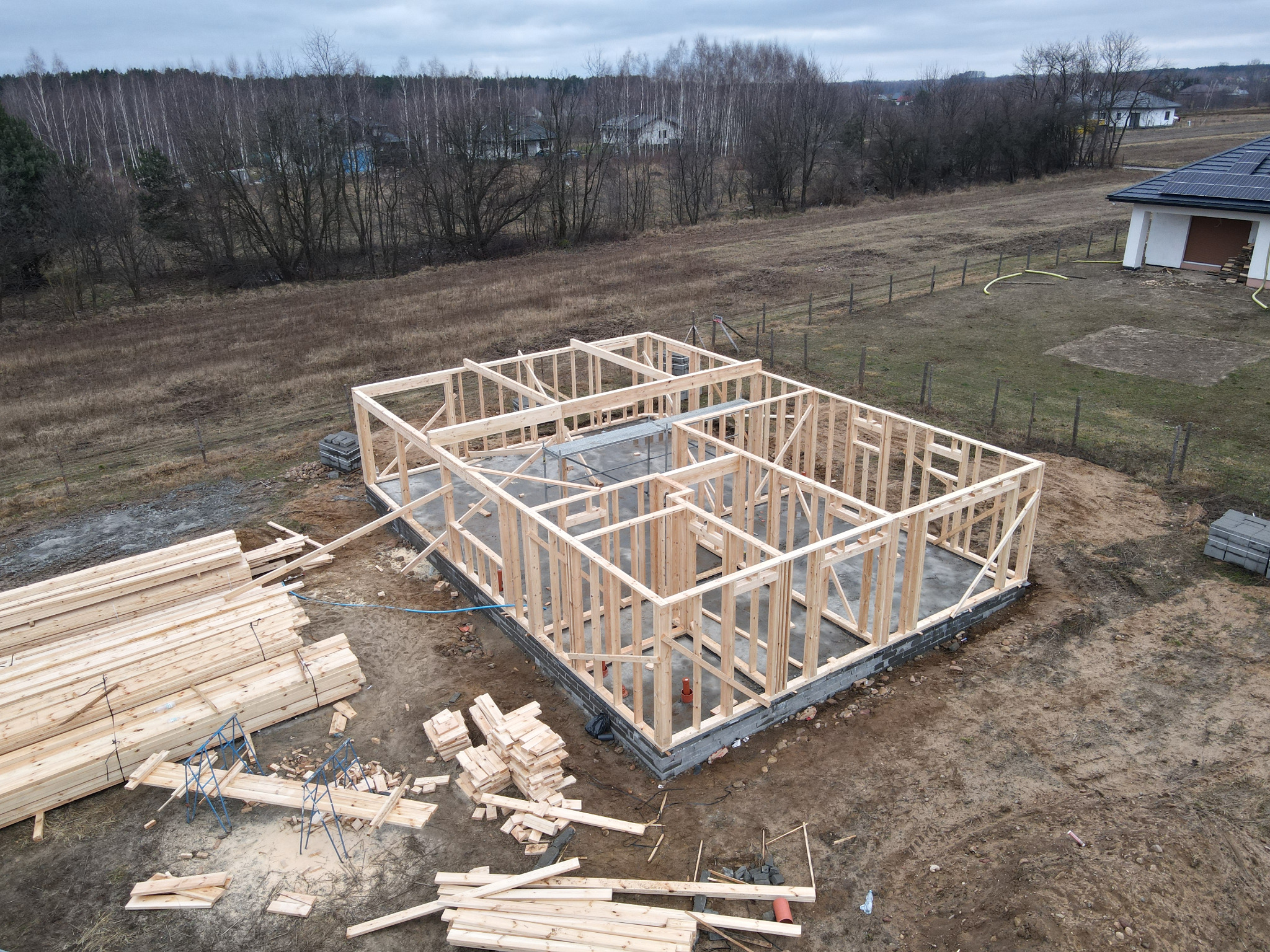 Wysoki kąt widzenia na szkieletową konstrukcję domu na betonowym fundamencie, z widocznym stosem drewna i elementami szalunku w tle, na tle brązowej trawy i drzew bezlistnych.