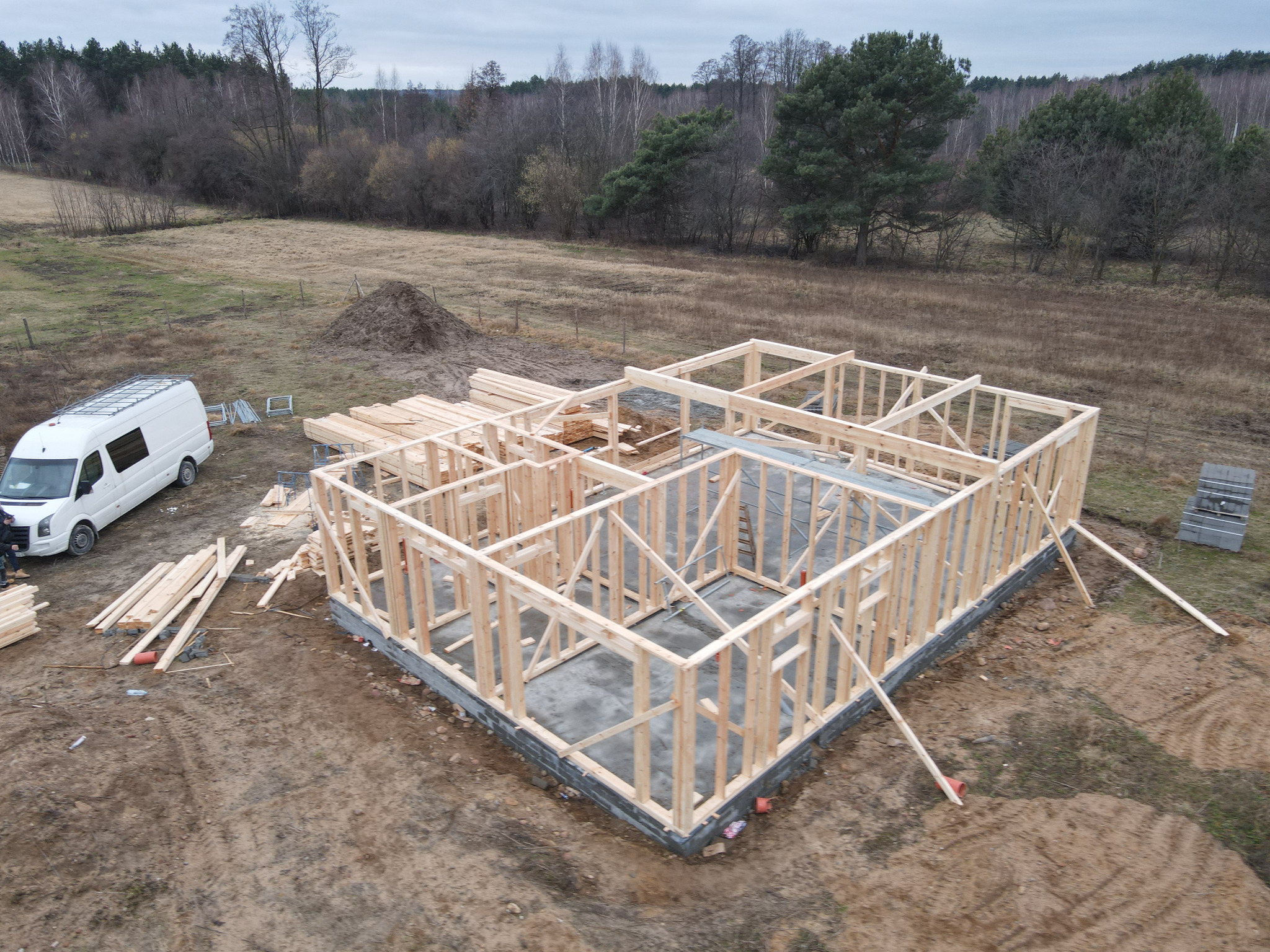 Wznoszenie konstrukcji domu z drewna na betonowym fundamencie, widok z góry, na placu budowy biały bus transportowy z drabinką na dachu, w tle las i sterta ziemi.