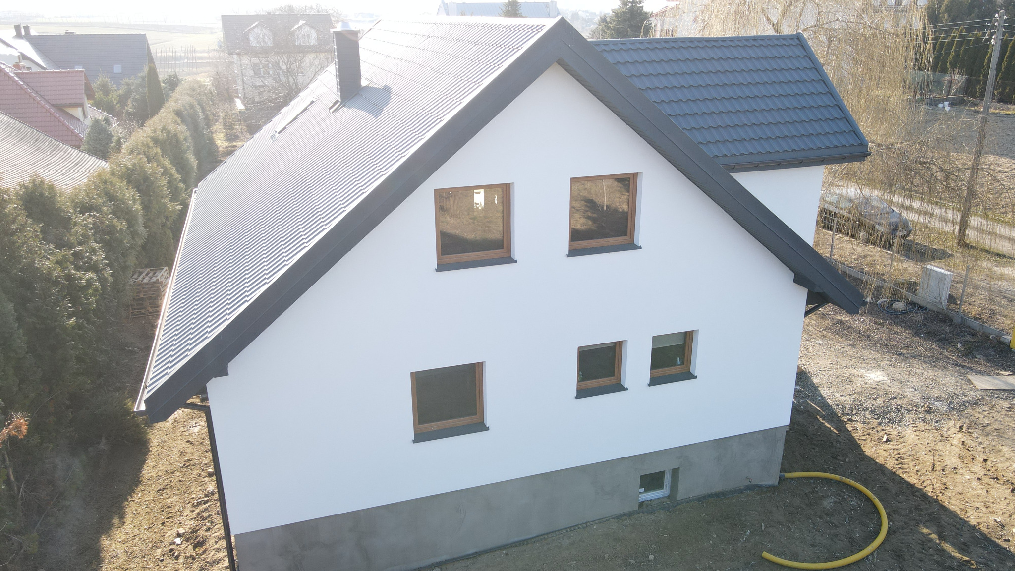 Nowoczesny, dwukondygnacyjny dom z dachem z blachodachówki w kolorze grafitowym, białymi ścianami i brązowymi oknami, widoczny z góry, z żółtą rurą drenażową przy fundamencie.