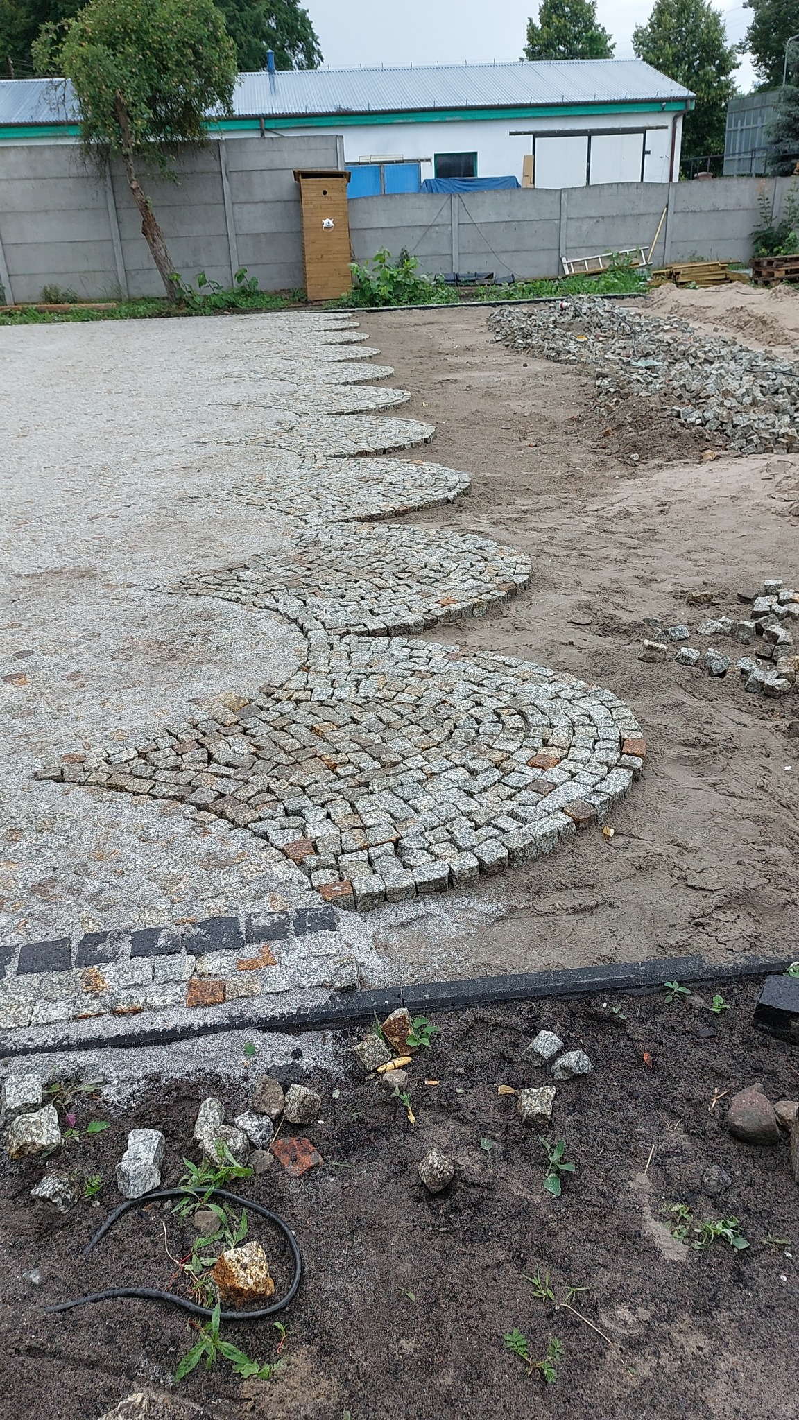 Wężykowaty wzór z jasnoszarej kostki granitowej w trakcie układania, widoczne przygotowane podłoże i fragmenty kostki do uzupełnienia, w tle budynek i betonowy płot.