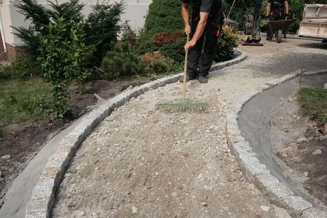 Przygotowanie podłoża pod ścieżkę z kostki granitowej: pracownik wyrównuje piasek grabiami, widoczne obrzeża z kostki granitowej i częściowo przygotowane korytko z betonu.