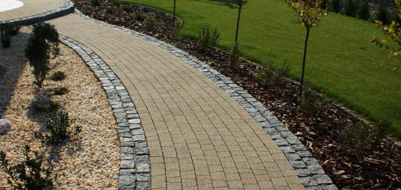 Zakrzywiony chodnik z beżowej kostki brukowej obramowany szarą kostką granitową, prowadzący przez zadbany ogród z trawnikiem, krzewami i korą.