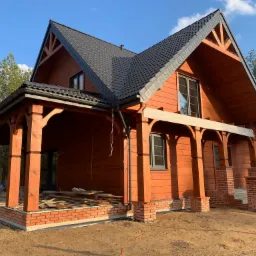 Nowoczesny dom z drewna z ciemną dachówką, widoczne elementy konstrukcyjne i fragment fundamentów w trakcie budowy.