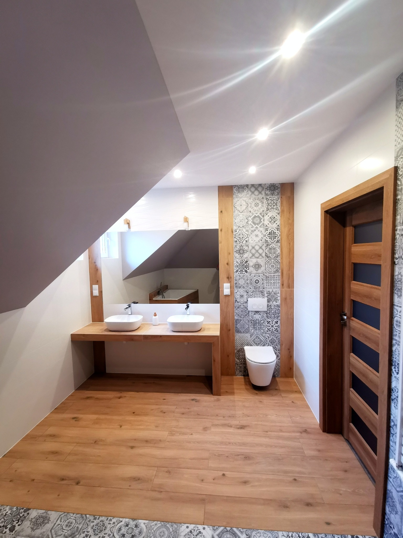 Nowoczesna łazienka na poddaszu z drewnopodobną podłogą, umywalkami nablatowymi, toaletą wiszącą i dekoracyjnymi płytkami za toaletą.