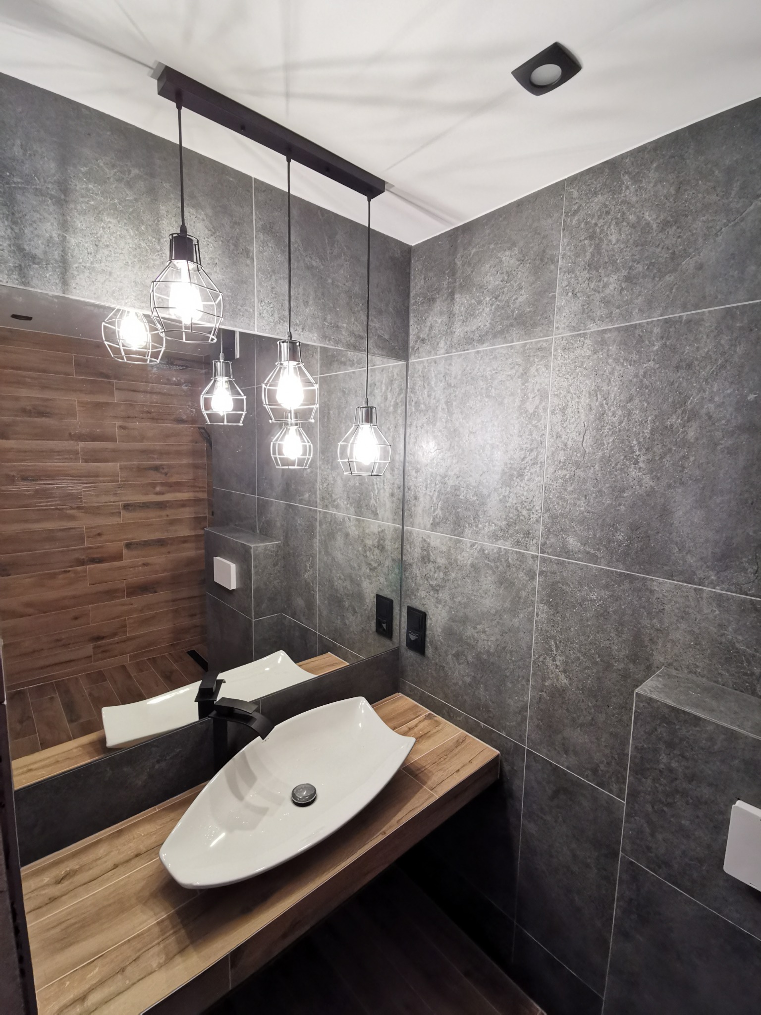 Nowoczesna łazienka z szarymi płytkami imitującymi kamień, drewnopodobnymi płytkami na ścianie prysznica, podwieszaną szafką z blatem z jasnego drewna i owalną białą umywalką oraz wiszącymi lampami...