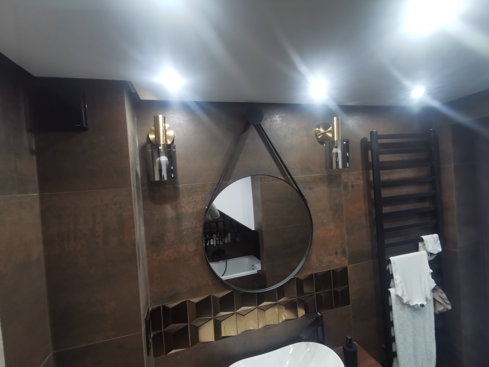 Nowoczesna łazienka z brązowymi płytkami, okrągłym lustrem na pasku, złotymi kinkietami i grzejnikiem drabinkowym z białymi ręcznikami.