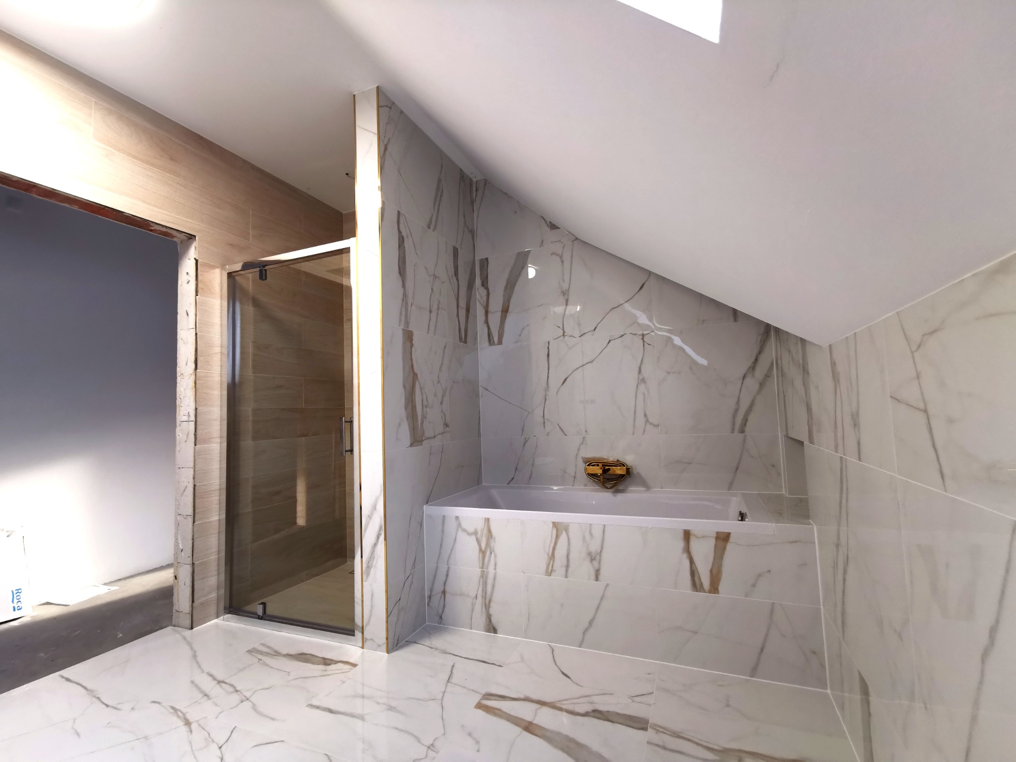 Nowoczesna łazienka na poddaszu z marmurowymi płytkami na podłodze i ścianach, kabina prysznicowa ze szklanymi drzwiami i złota armatura nad wanną.