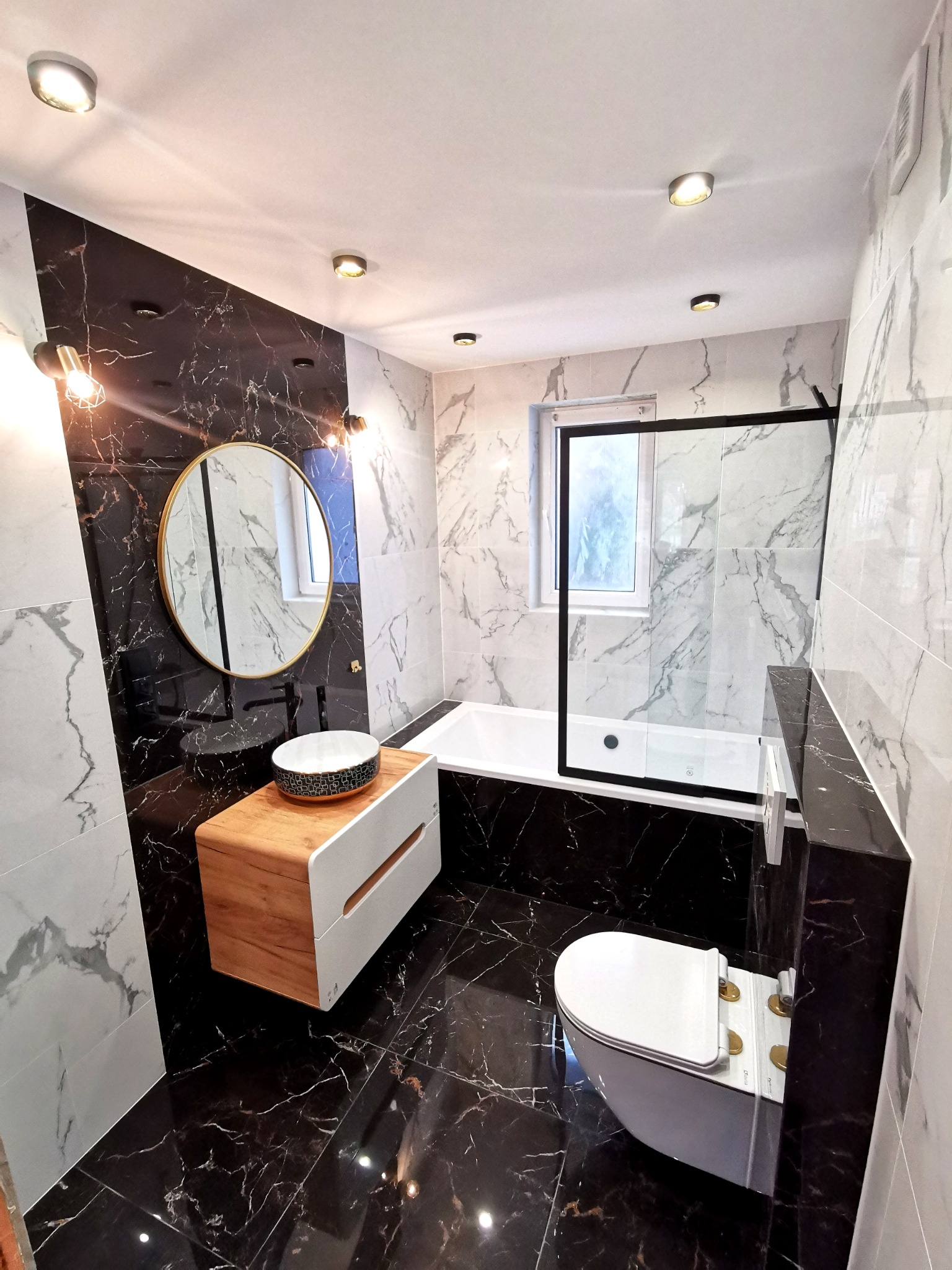 Nowoczesna łazienka z czarno-białymi marmurowymi płytkami, okrągłym lustrem w złotej ramie, umywalką nablatową na drewnianej szafce, białą wanną z czarną szybą i podwieszaną toaletą.