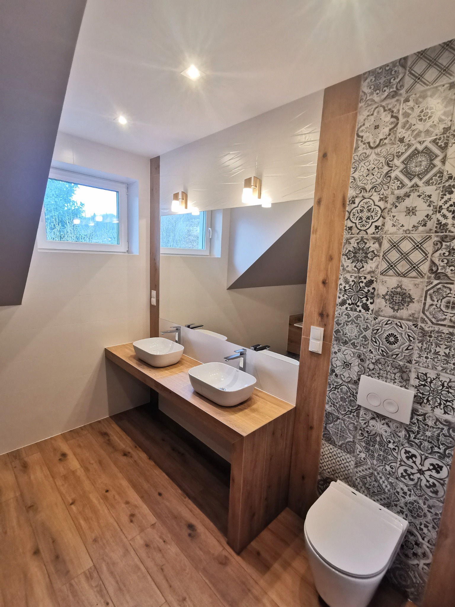 Nowoczesna łazienka na poddaszu z drewnianą podłogą, blatem z umywalkami nablatowymi i ścianą wykończoną wzorzystymi płytkami w stylu patchworkowym, oświetlona punktowymi lampami.
